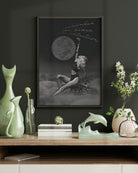 female energy dream artwork in a frame of a woman holding a moon over clouds on a dark wall with decorative items below, 