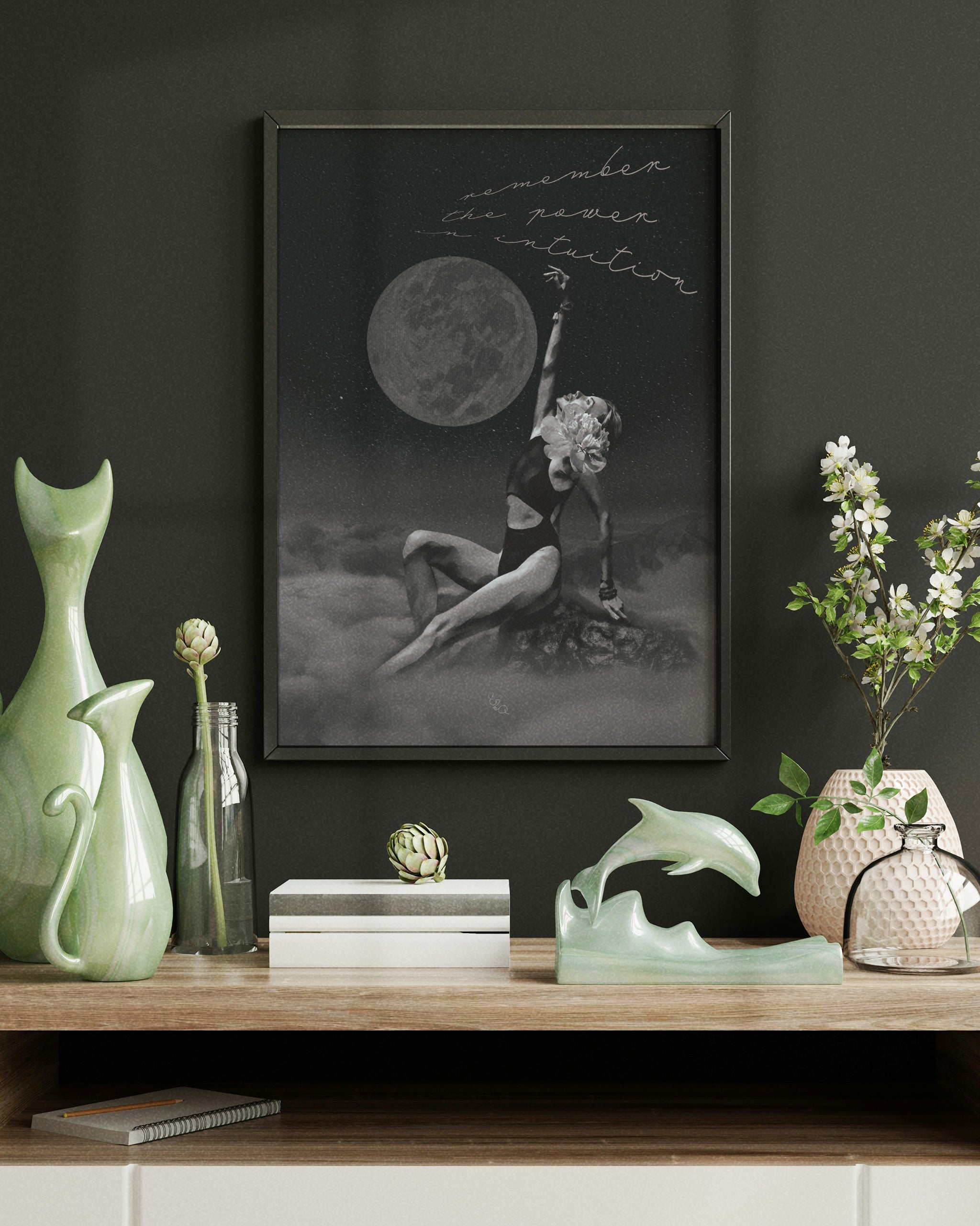 female energy dream artwork in a frame of a woman holding a moon over clouds on a dark wall with decorative items below, 
